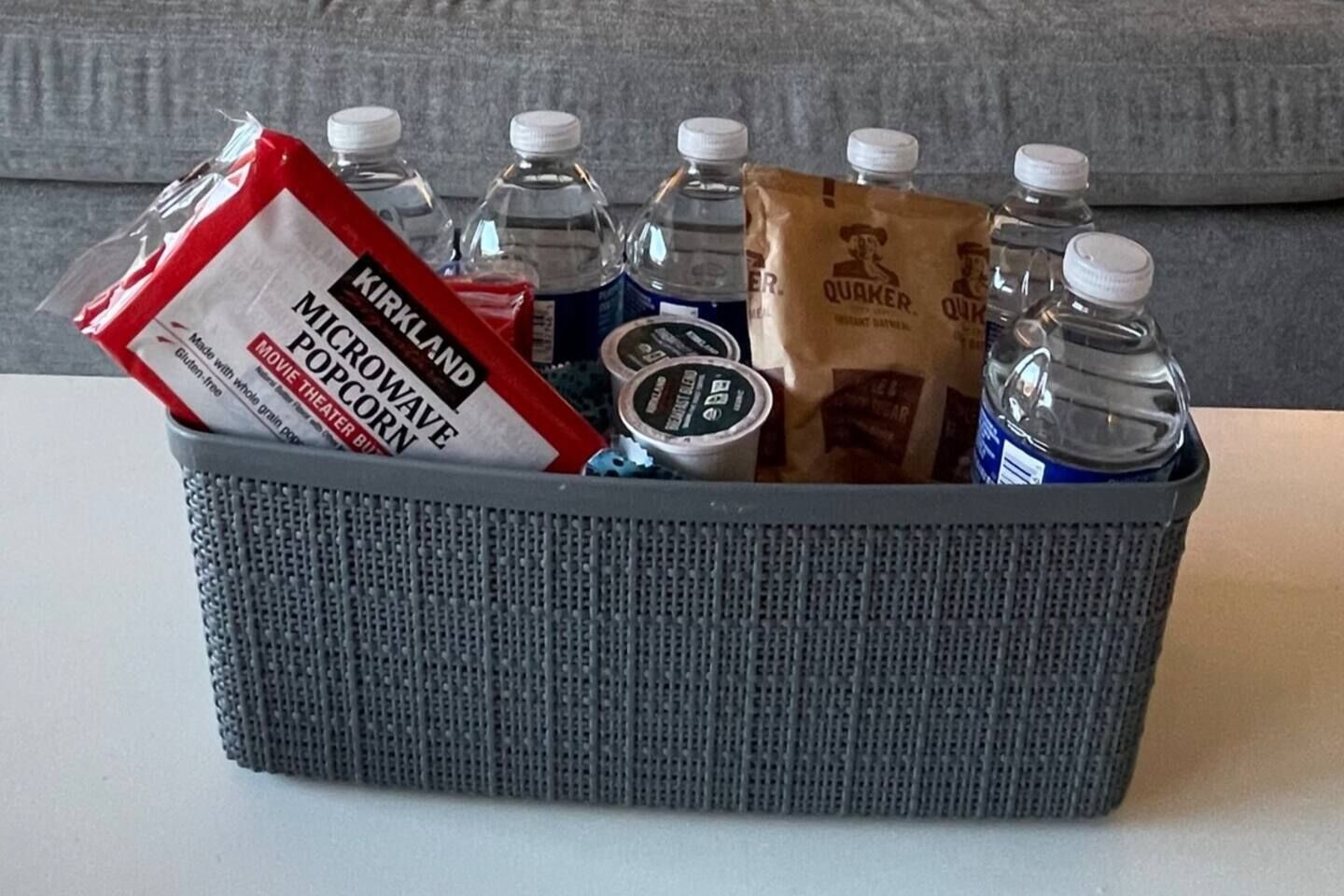 A gray basket containing Kirkland microwave popcorn, Quaker Oats, water bottles, and a container of butter.