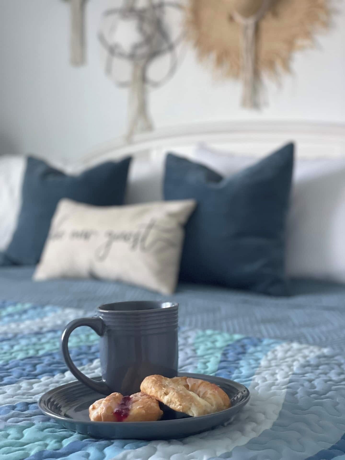 A bed with a blue quilt, a gray mug, and a plate with pastries.