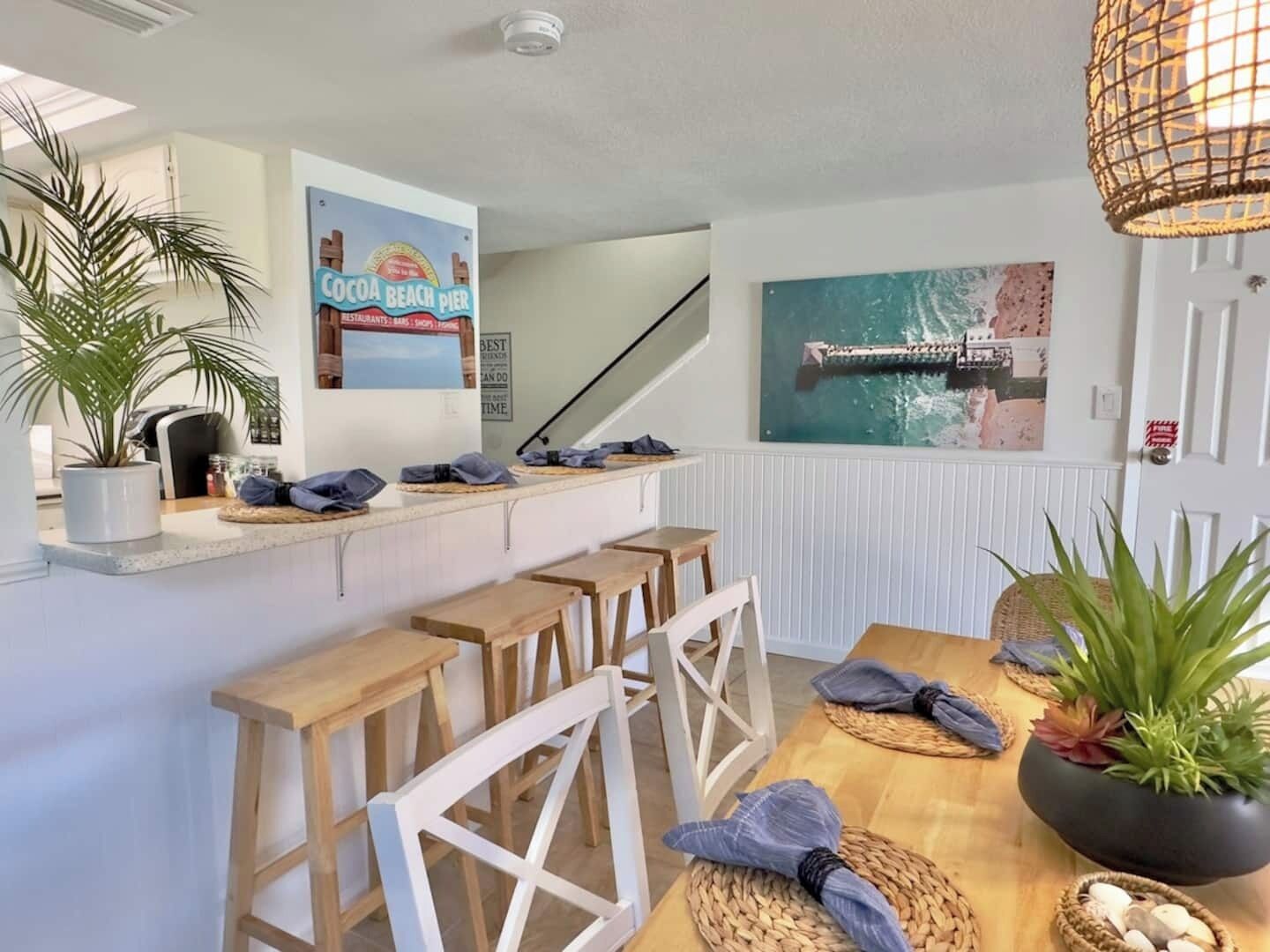 A kitchen area with a counter, stools, and a dining table. There is a sign on the wall and a painting above the door.