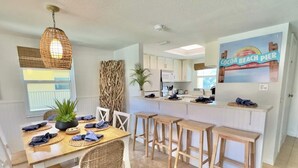 A modern kitchen with white cabinets, a central island with stools, and a dining area with wicker chairs and a table set for four. - Beachfront with heated pool, 60 seconds to beach (Cocoa Beach)