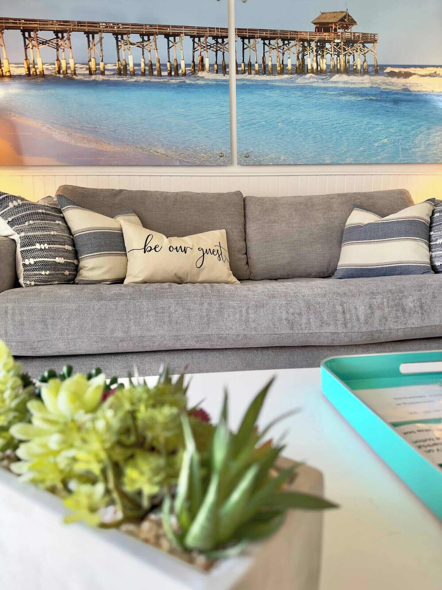 A grey sofa with a cushion that reads 'be our guest' in front of a wall with a painting of a pier and ocean.