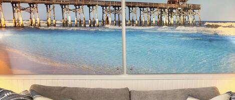 A grey sofa with a cushion that reads 'be our guest' in front of a wall with a painting of a pier and ocean.