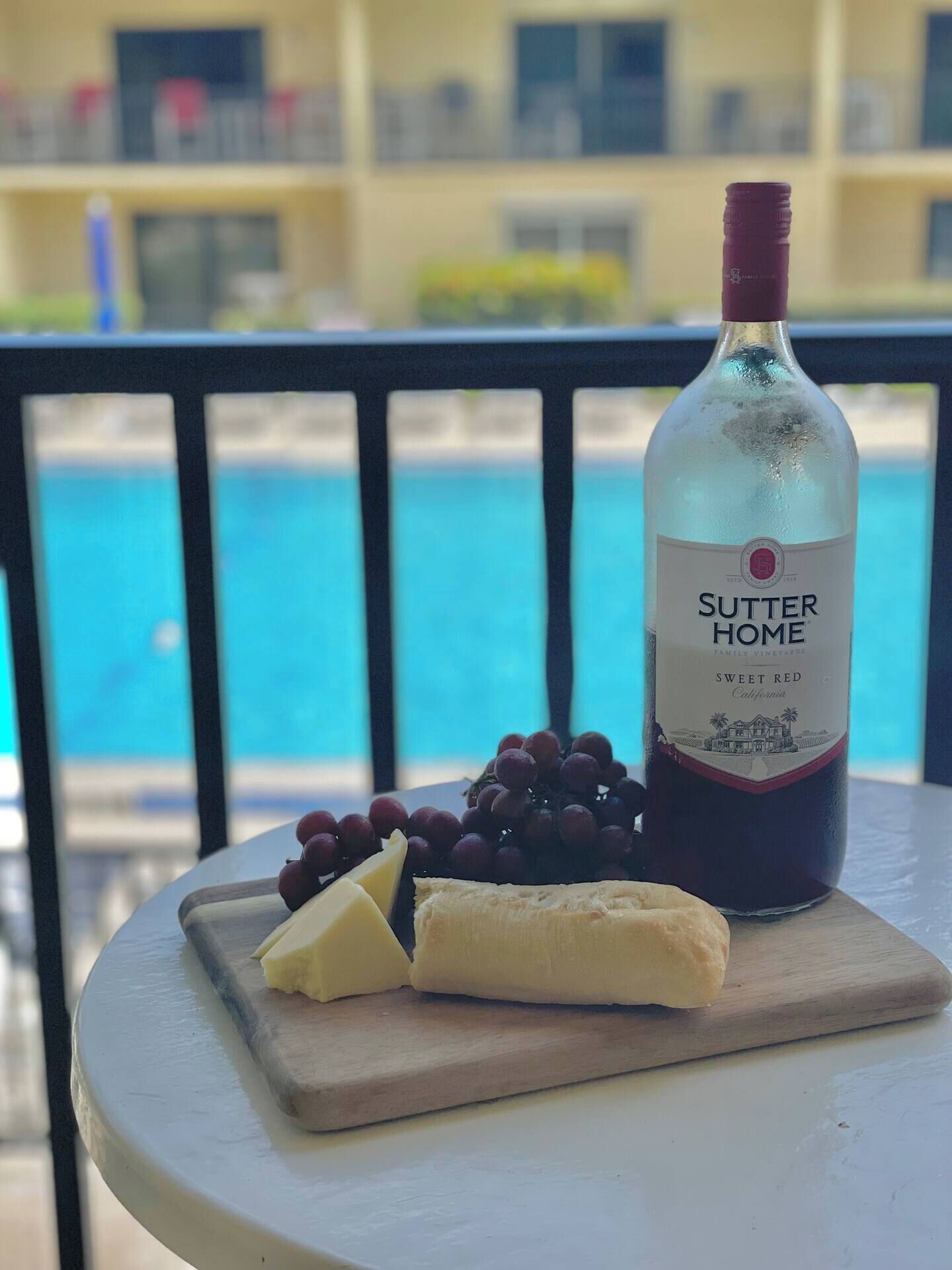 A bottle of Sutter Home Sweet Red wine, a wooden cutting board with cheese and grapes, and a pool in the background.