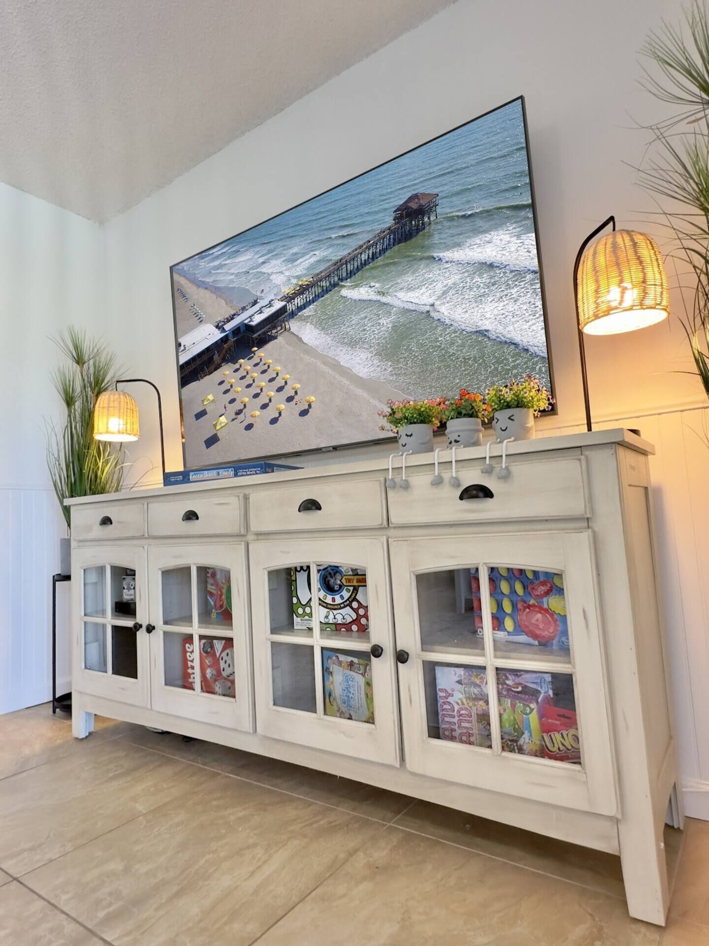 A white cabinet with glass doors displaying board games, a large TV mounted above displaying a pier and beach scene, and two floor lamps on either side.