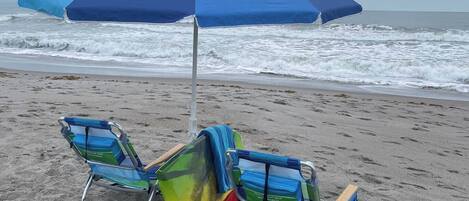 A beach with two beach chairs and a blue umbrella.