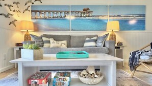 A living room with a grey sofa, a white coffee table, and a painting of a pier and ocean. There are two lamps, a clock, and a basket of seashells on the coffee table. - Beachfront with heated pool, 60 seconds to beach (Cocoa Beach)