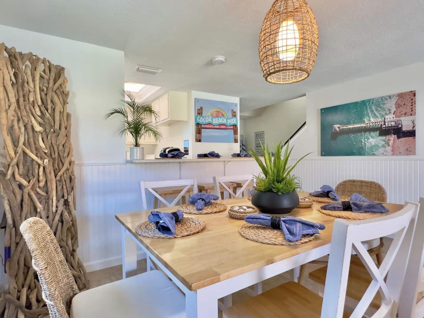 Un comedor con mesa de madera, sillas blancas, individuales tejidos y una lámpara colgante.