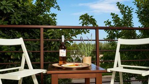 Ein Holztisch mit zwei weißen Stühlen, einer Weinflasche und zwei Weingläsern auf einem Balkon mit Metallgeländer und Grünpflanzen im Hintergrund.
