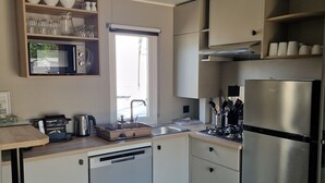 A modern kitchen with a stainless steel refrigerator, a built-in oven, and a coffee machine. There is a window with a view of the outdoors, and a shelf with bowls and glasses.