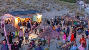 A beachside restaurant with outdoor seating, surrounded by sand dunes and a wooden deck.