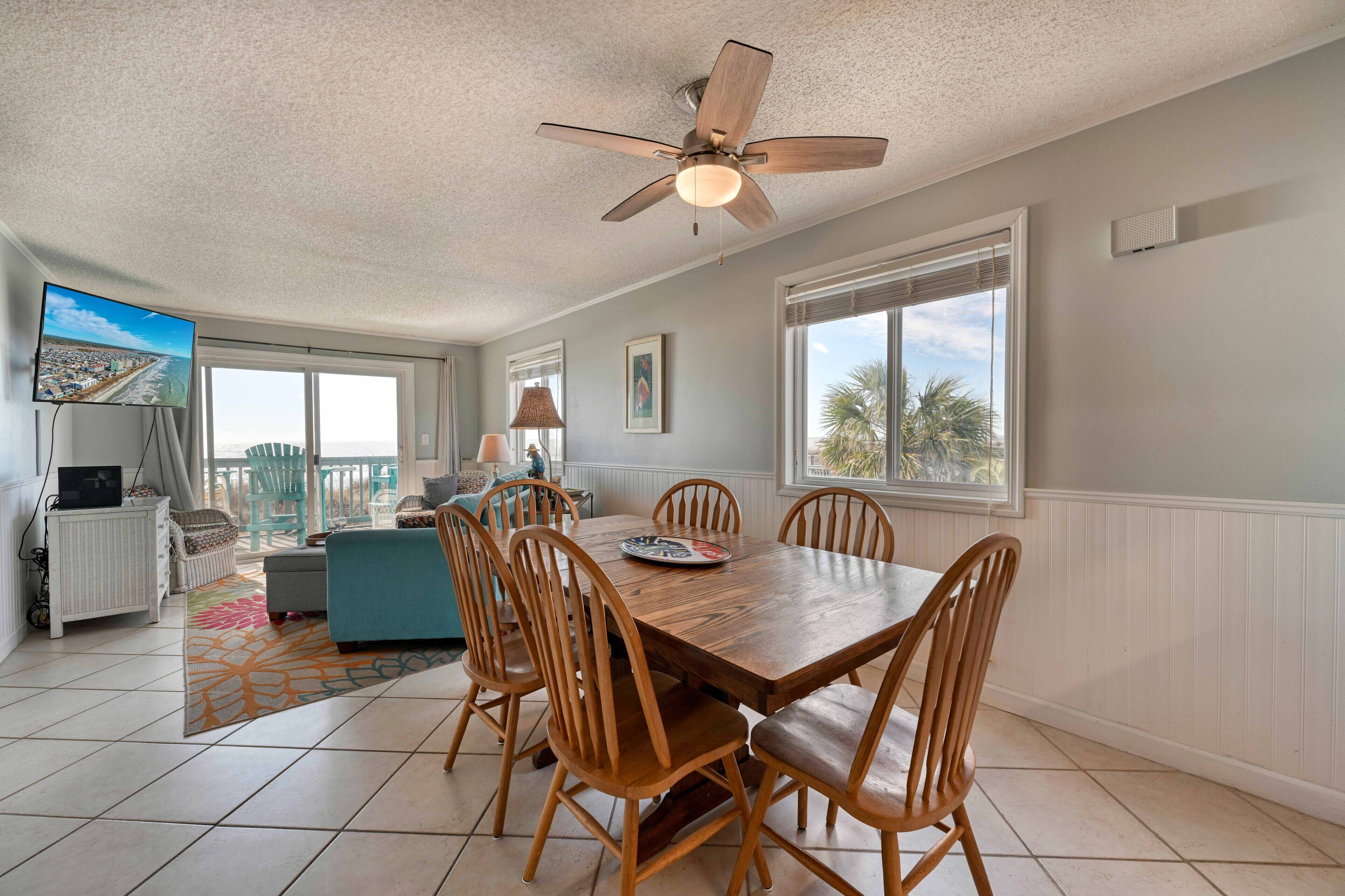 A spacious living room with a wooden dining table, comfortable seating, a ceiling fan, and a television on a stand.
