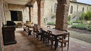 Un patio exterior con una larga mesa de madera y bancos, rodeado de pilares de ladrillo y vistas a un edificio histórico.