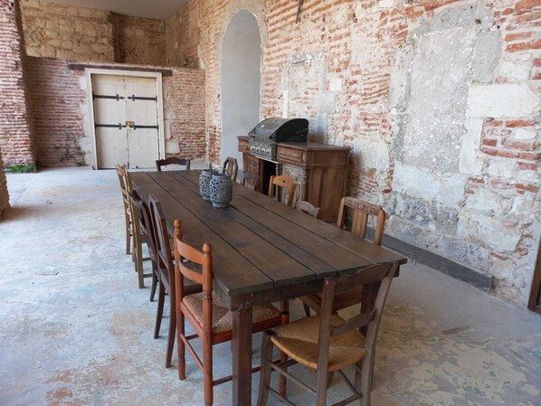 Un coin repas rustique avec une longue table et des chaises en bois, un mur en briques et un petit barbecue.