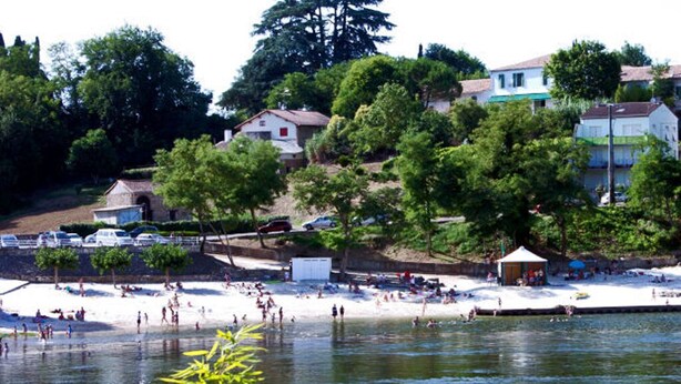 Una playa con gente nadando y tomando el sol, rodeada de árboles y casas.