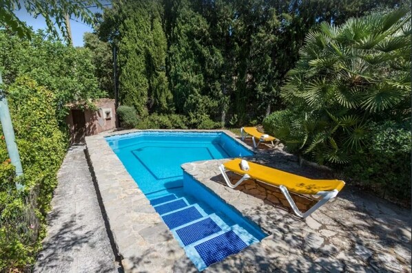 Una piscina rectangular con borde de piedra, dos tumbonas amarillas y un tapete de seguridad azul.