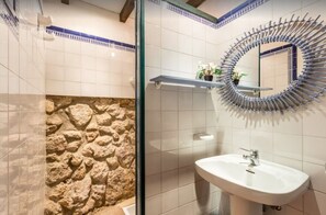 Un baño con pared de piedra, cabina de ducha de vidrio, lavamanos blanco y espejo circular.