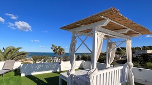 Un quiosco con barandas blancas y techo de madera, rodeado de sillas de lounge blancas y césped artificial, con vista a una playa y al océano.