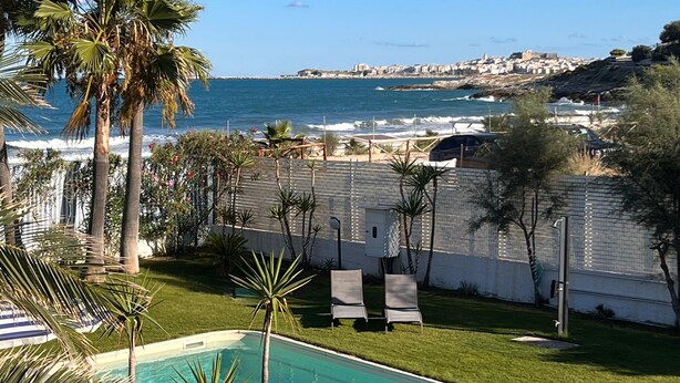 Una piscina con agua cristalina, rodeada de palmeras y vista al mar y edificios a lo lejos.