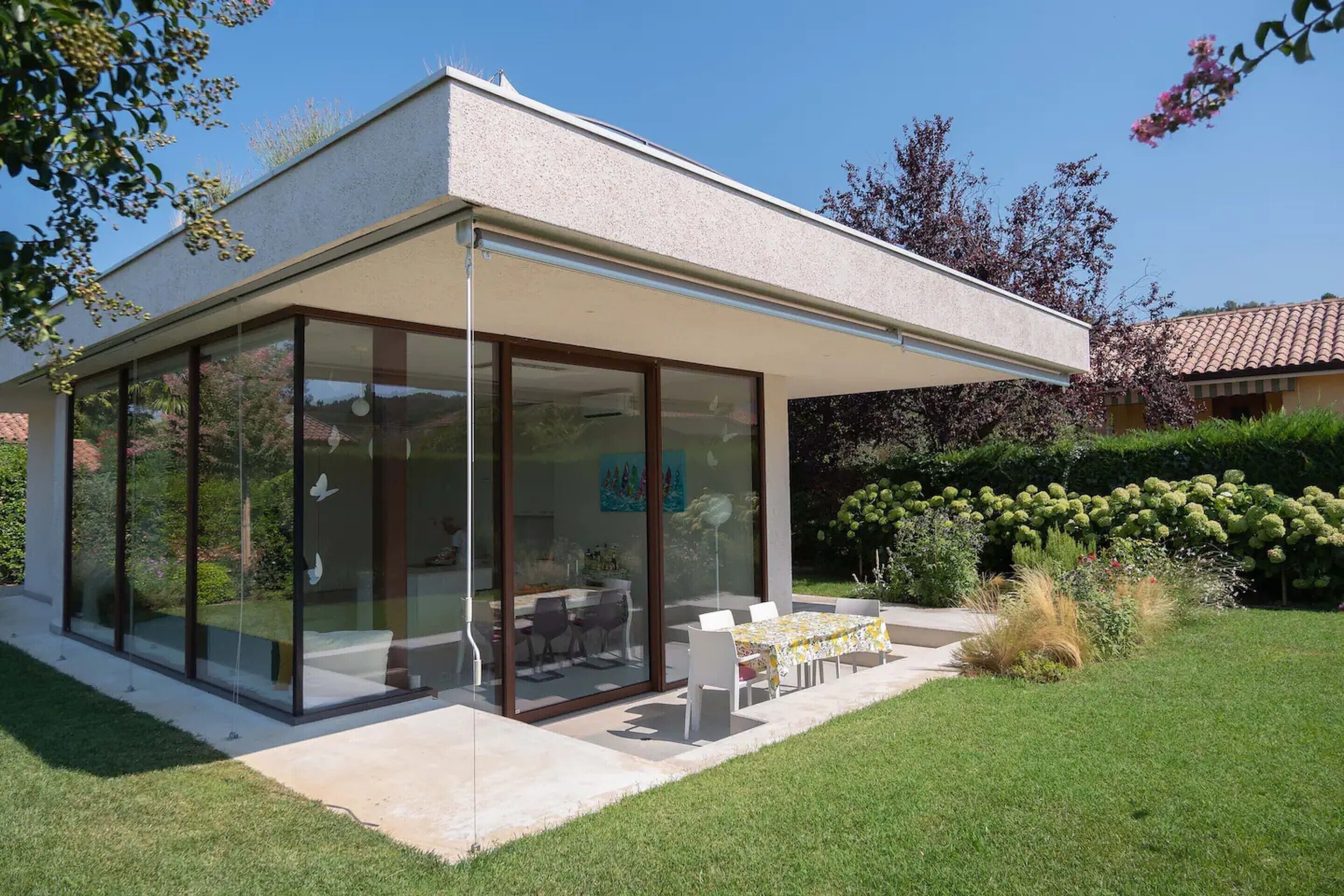 Una casa moderna con un patio envidriado, un comedor con sillas y un jardín con diversas plantas.