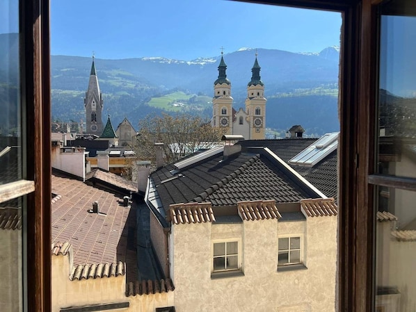 A view of a town with traditional architecture, including a church with a clock tower and two spires, through a window with a wooden frame.