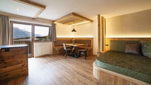 Une pièce avec un bureau en bois, un lit, une fenêtre donnant sur un beau paysage et un plafond avec des poutres apparentes.