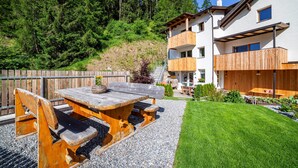 Une table et des bancs en bois installés à l’extérieur, un bâtiment blanc doté de balcons et une colline boisée.