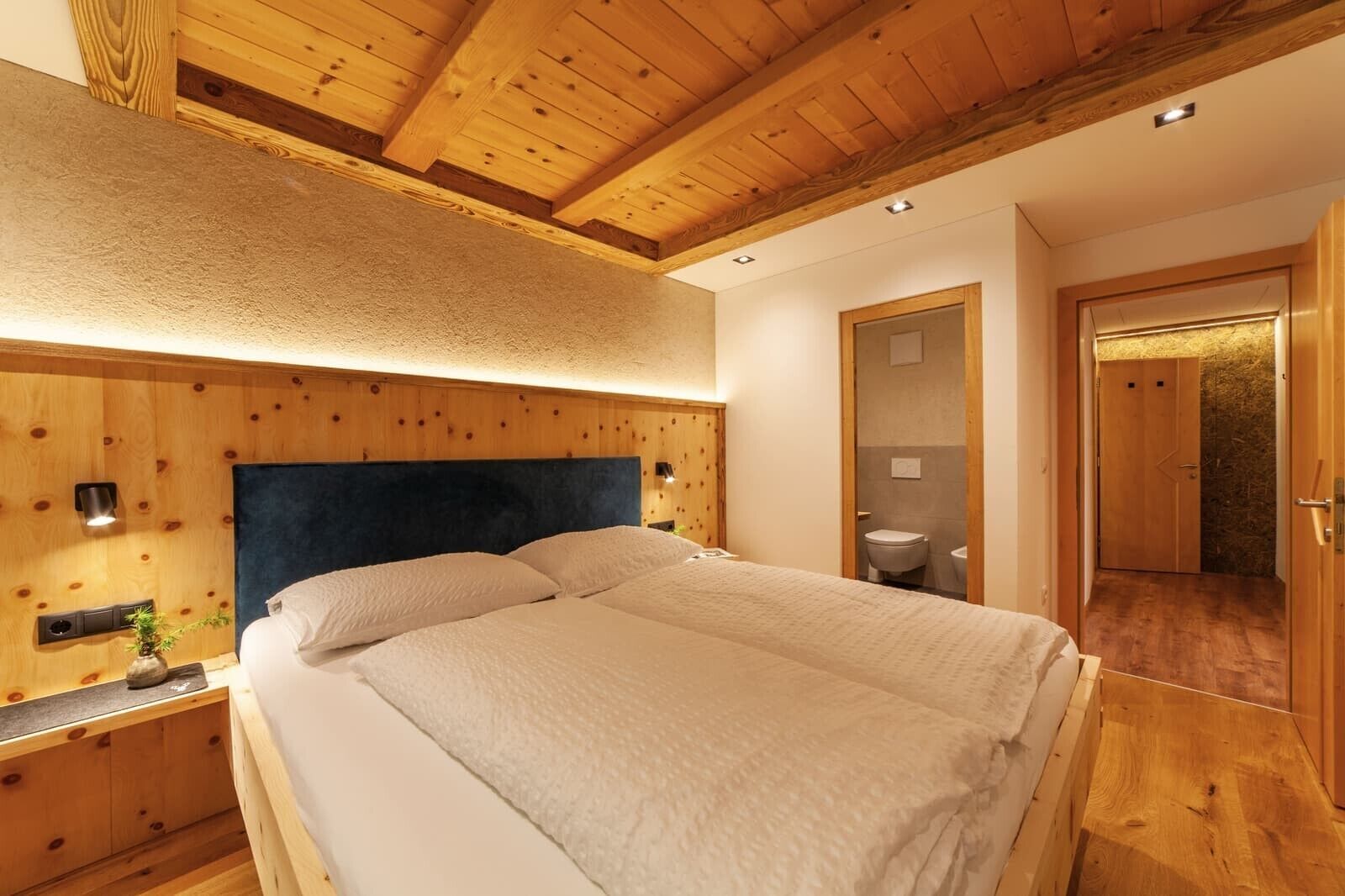 Ein Schlafzimmer mit Holzwandverkleidung, einem Bett, Nachttischen und einem durch eine offene Tür einsehbaren Badezimmer.