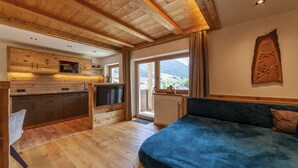 Ein Zimmer mit Holzbalkendecke, einem Bett, einem Fernseher, einer Küchenzeile und einem großen Fenster mit Bergblick.