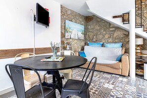 A dining area with a round table, chairs, a TV mounted on the wall, a painting, a sofa with cushions, and a stone wall. - Holiday Home 'Casa Azul' with Mountain View, Shared Pool and Wi-Fi (Moledo)