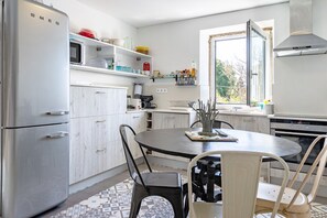 A modern kitchen with white cabinetry, a stainless steel refrigerator, a round dining table with chairs, and a window providing natural light. - Holiday Home 'Casa Azul' with Mountain View, Shared Pool and Wi-Fi (Moledo)