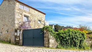 A stone building with a closed dark gate, a cobblestone street, and a lush green hedge. - Holiday Home 'Casa Azul' with Mountain View, Shared Pool and Wi-Fi (Moledo)