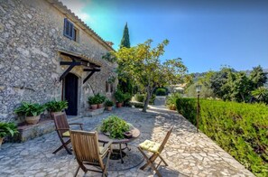 Un bâtiment en pierre doté d’une porte et de fenêtres, entouré de plantes en pot et d’une terrasse pavée aménagée avec du mobilier d’extérieur.