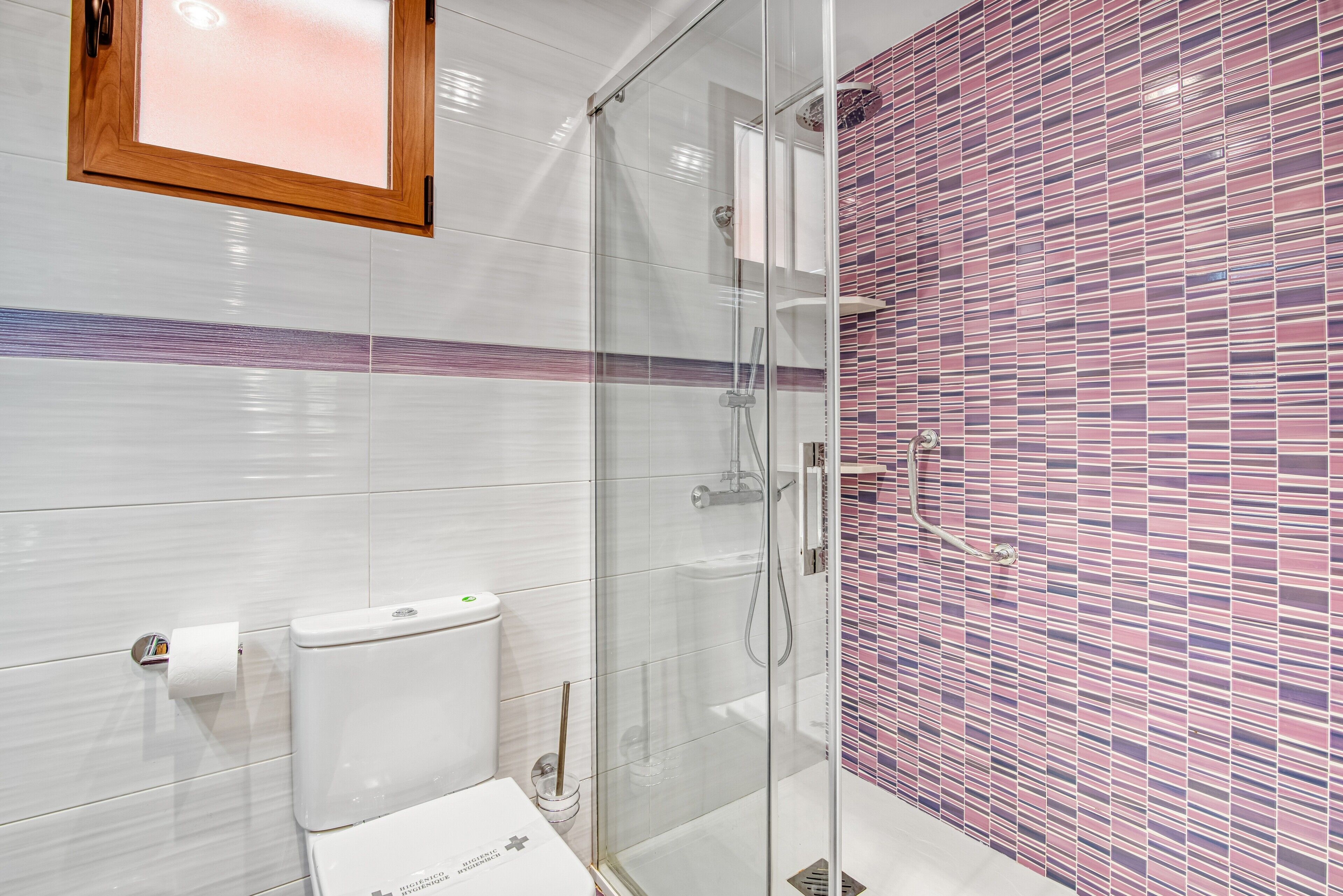 Un baño moderno con cabina de ducha de vidrio, inodoro y pared de azulejos con estampado en rosa y morado.