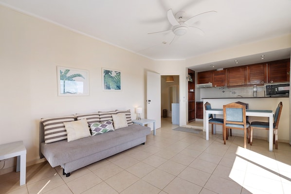 A living room with a sofa, a dining table with chairs, a ceiling fan, and a kitchen area with cabinets and appliances.