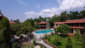 A pool surrounded by lush greenery and traditional buildings with red-tiled roofs. - Safari Club (Sauraha)