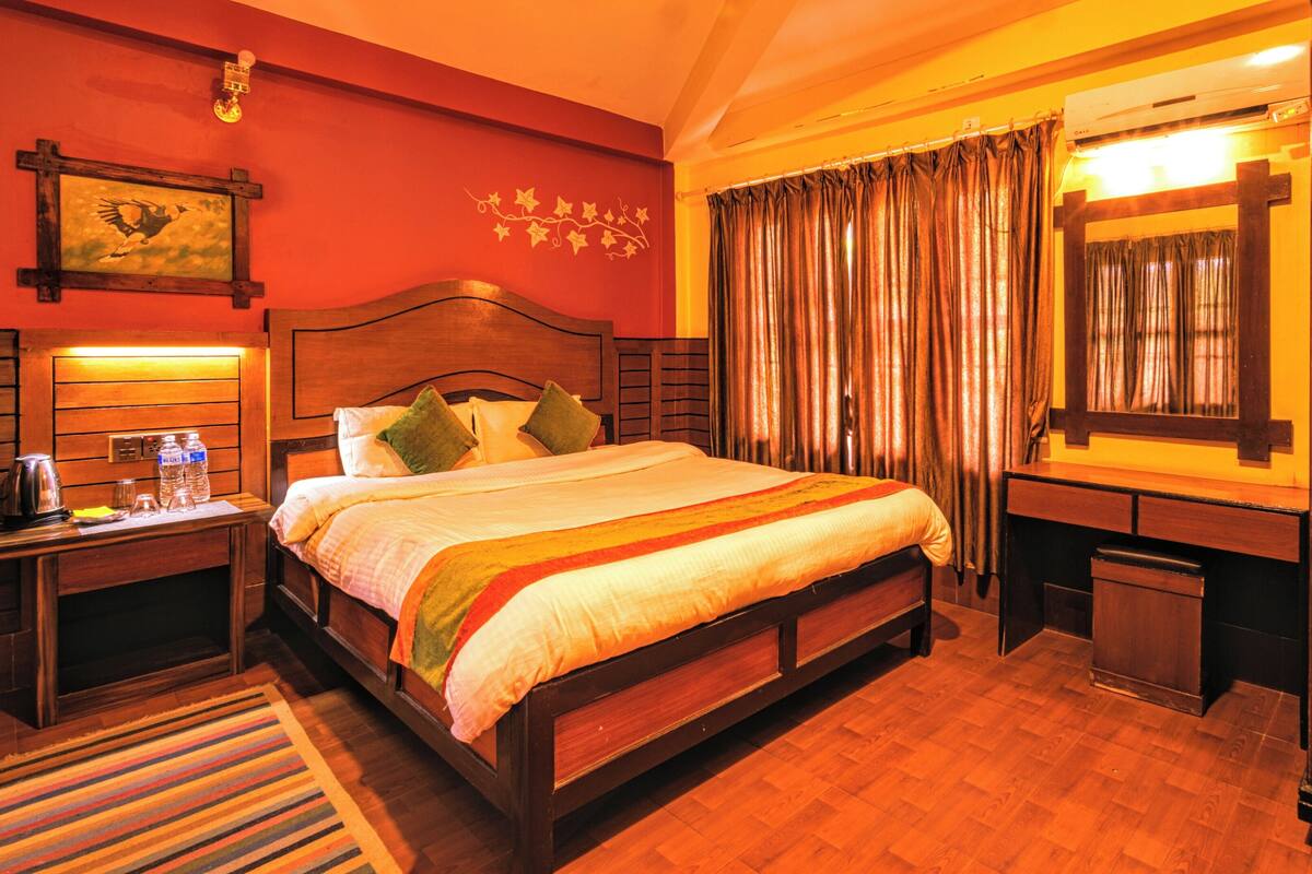 A hotel room with a wooden bed, a nightstand with a kettle and water bottles, a mirror, and a window with curtains.