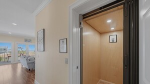 Een gang met een houten vloer, een grijze bank en een deur die leidt naar een kamer met een houten plafond.