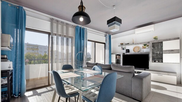 A modern living room with a glass dining table, blue chairs, a grey sofa, and a flat-screen TV. - Apartment "Refúgio Praia da Rocha" with Sea View, Balcony & Wi-Fi (Portimão)