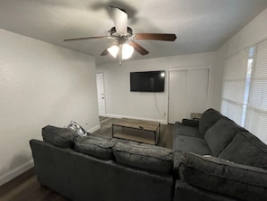 A living room with a sectional sofa, a coffee table, a ceiling fan, and a flat-screen TV mounted on the wall. - Spacious 3-Bedroom Home Convenient to Fort Sill (Lawton)