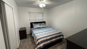 Un dormitorio con cama, mesita de noche, ventilador de techo y ventana con persianas.