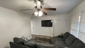 Una sala de estar con un sofá modular, una mesa de centro, un ventilador de techo y un televisor de pantalla plana montado en la pared.