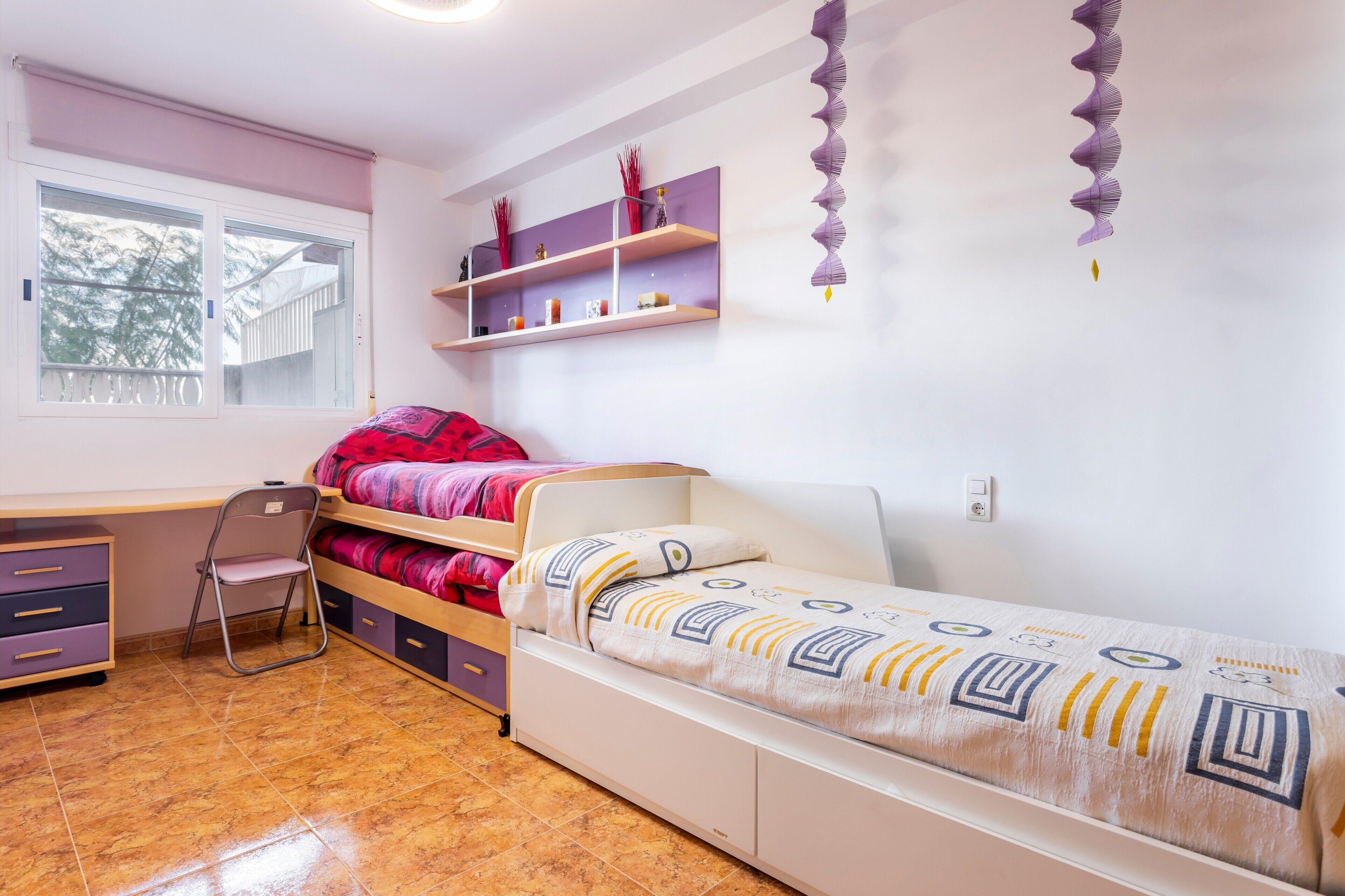 Een slaapkamer met een bed, een bureau en een stoel. Boven het bed zijn planken en er is een raam met jaloezieën.