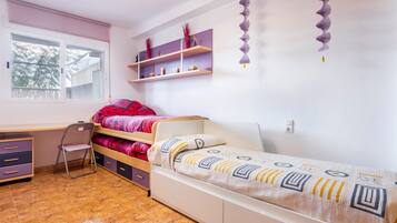 Een slaapkamer met een bed, een bureau en een stoel. Boven het bed zijn planken en er is een raam met jaloezieën.