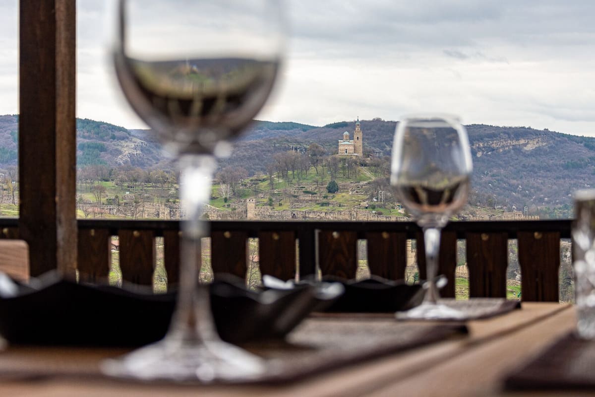 A table with two wine glasses and a glass of water, set against a backdrop of a mountainous landscape with a castle.