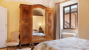 A bedroom with a bed, a wooden wardrobe, a striped armchair, and a window with a view of a brick building. - Apartment 'La Rocca di Efisio - Grignolino' with Balcony, Wi-Fi and Air Conditioning (Costigliole d'Asti)