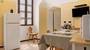 A compact kitchen with a refrigerator, a dining table with chairs, a flower vase, and a television mounted on the wall. - Apartment 'La Rocca di Efisio - Grignolino' with Balcony, Wi-Fi and Air Conditioning (Costigliole d'Asti)