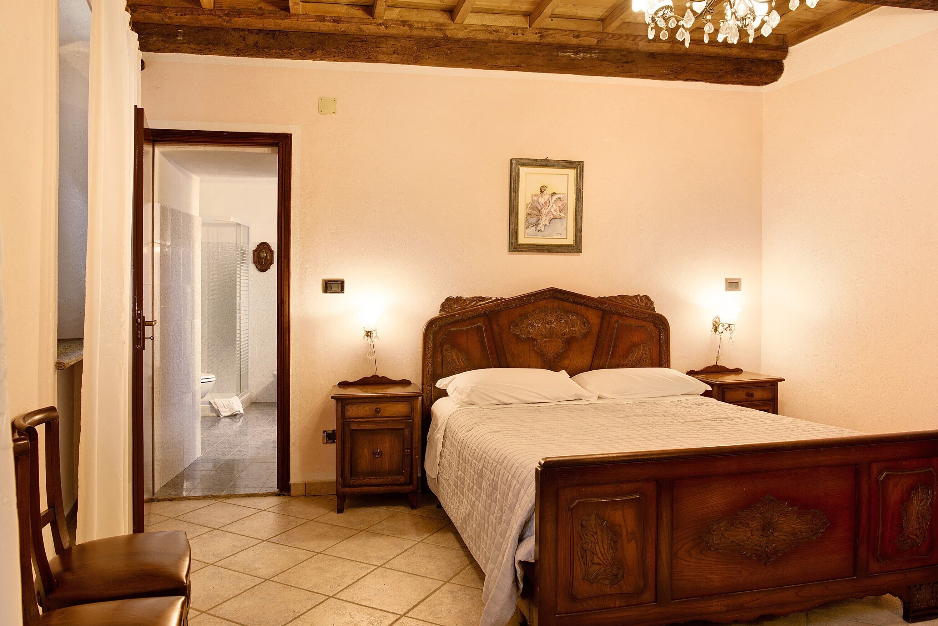A bedroom with a wooden bed, two bedside tables, a chair, and a door leading to a bathroom.