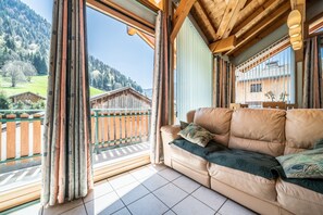 A room with a leather sofa, a view of a wooden cabin, and a balcony with a wooden railing.