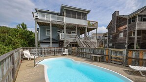 En swimmingpool med træterrasse, omgivet af et træhegn. Liggestole og en trappe, der fører op til en altan på anden sal.