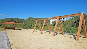 En gungställning i trä på en sandstrand, med ett trädäck och en lusthus i bakgrunden.